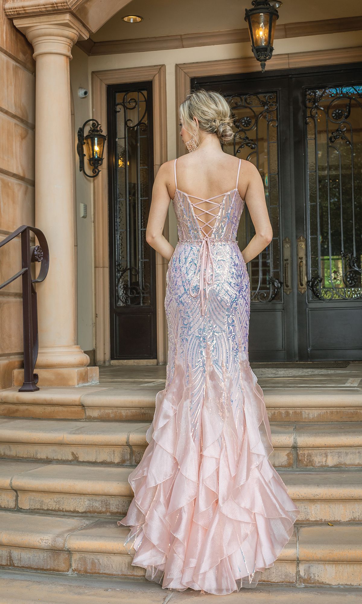 A woman in the Long Sequin Mermaid Prom Dress with Ruffles 4221, featuring pale pink sequins and ruffled details, stands on stone steps with her hair in an updo, facing ornate double doors.